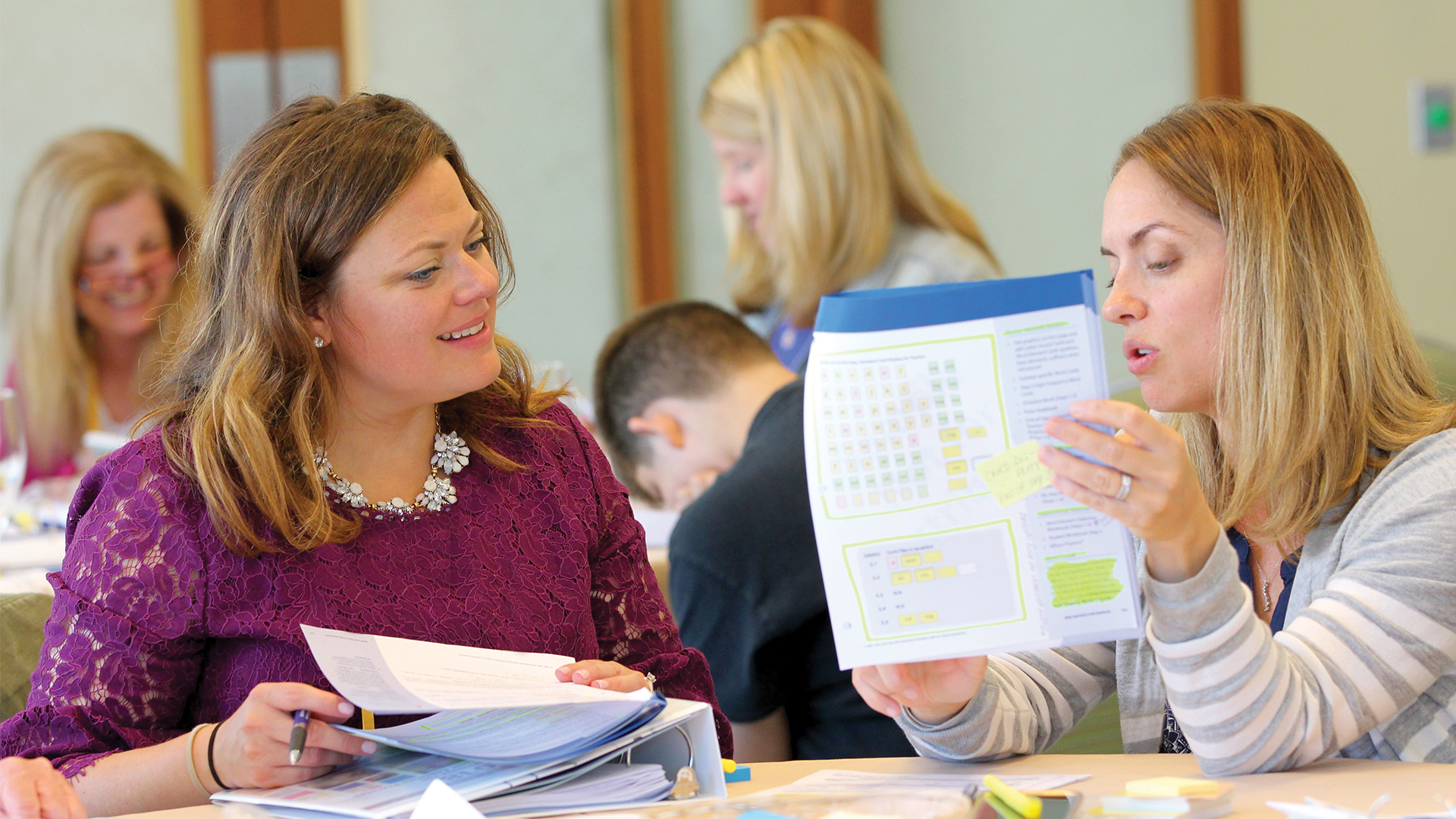Two women discuss training materials, with other attendees in background