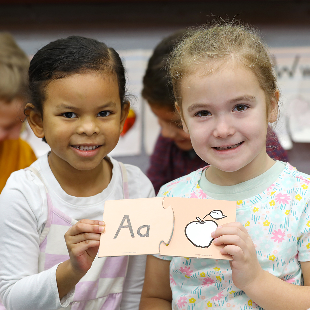 Two young girls hold an alphabet card with 'Aa' and an apple illustration.