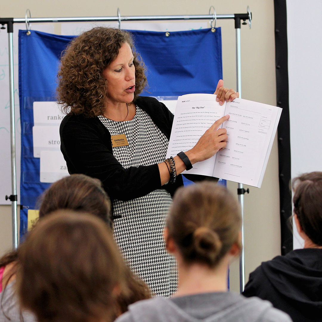 Teacher with curly hair points to a page in a book for students