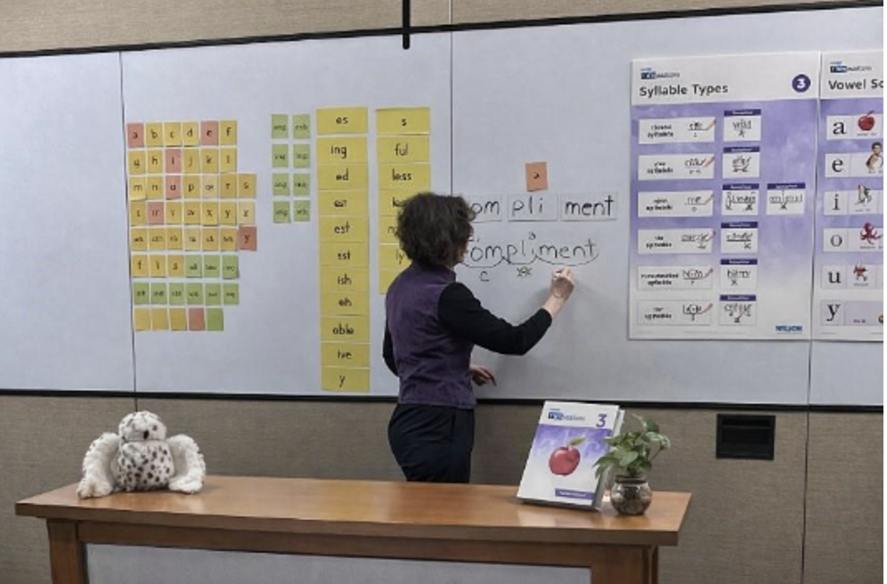 Fundations® workshop: teacher writes "compliment" on whiteboard with syllable charts