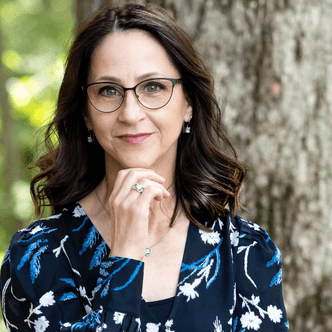 Woman with glasses and dark hair wearing a floral dress, thoughtful pose.