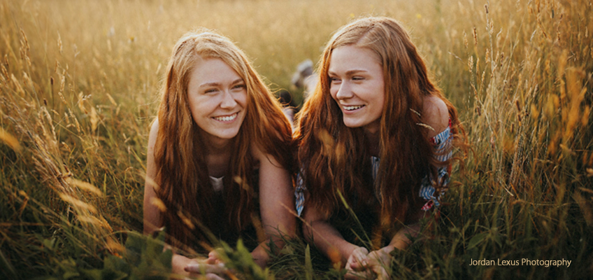 Two young women with red hair lying in a field, smiling and looking at each other.
