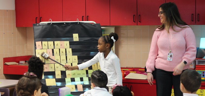 Teacher and students at a learning chart with letters and words, discussing NAEP scores.