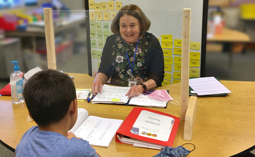 Teacher and student at desk with protective shield, focusing on reading.