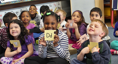 Children in a classroom holding letter cards, including 'D' and 'E', from the Fundations Pre-K Activity Set.
